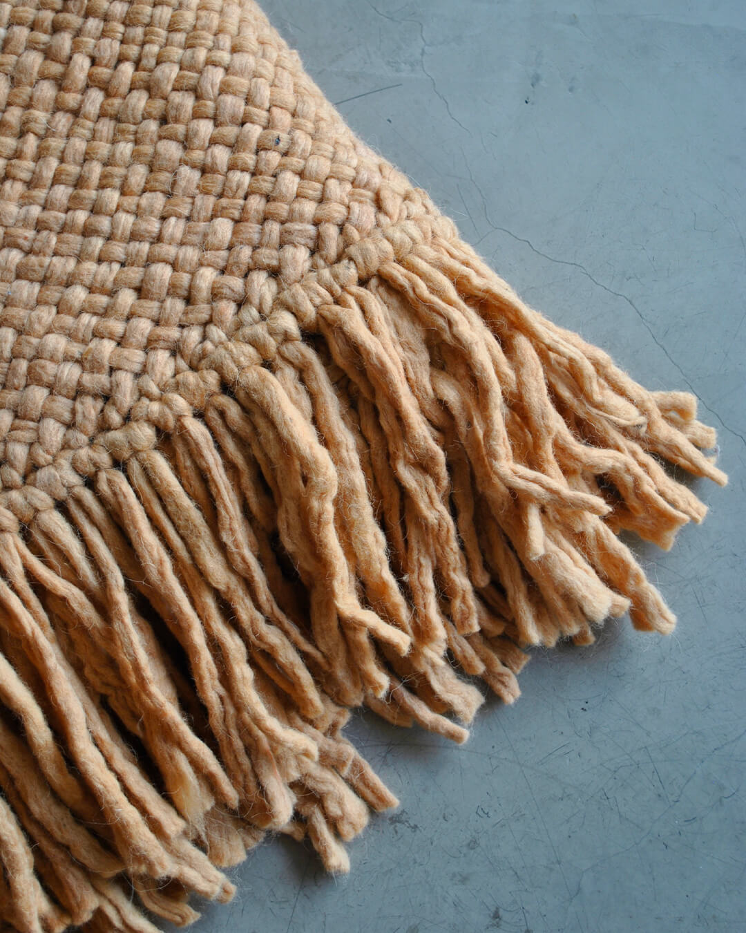 Camel-colored wool throw with handmade fringes on a concrete floor.