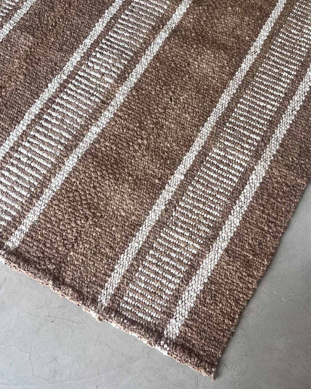 Handwoven rug detail in wood and natural tones, showing comb-style stripes and dense wool texture on a concrete floor.
